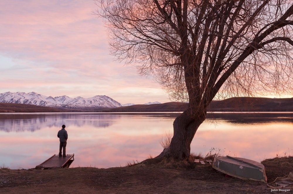 A man stands on a jetty looking out at a colourful dawn over Lake Alexandrina. The southern end of Lake Alexandrina has a collection of tiny holiday cottages.
